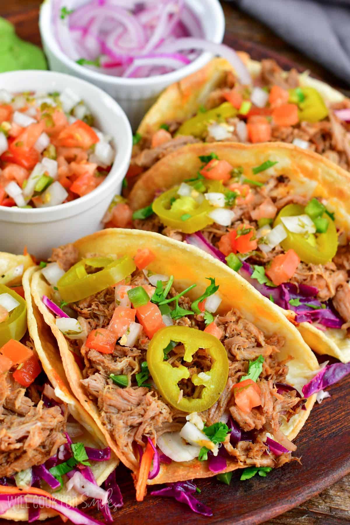 Four pulled pork tacos on a wooden plate with pickled jalapenos, diced tomato and Pico de Gallo on top.