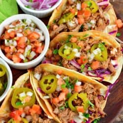 Four pulled pork tacos on a wooden plate with red onion, Pico de Gallo and pickled jalapenos in small bowls.