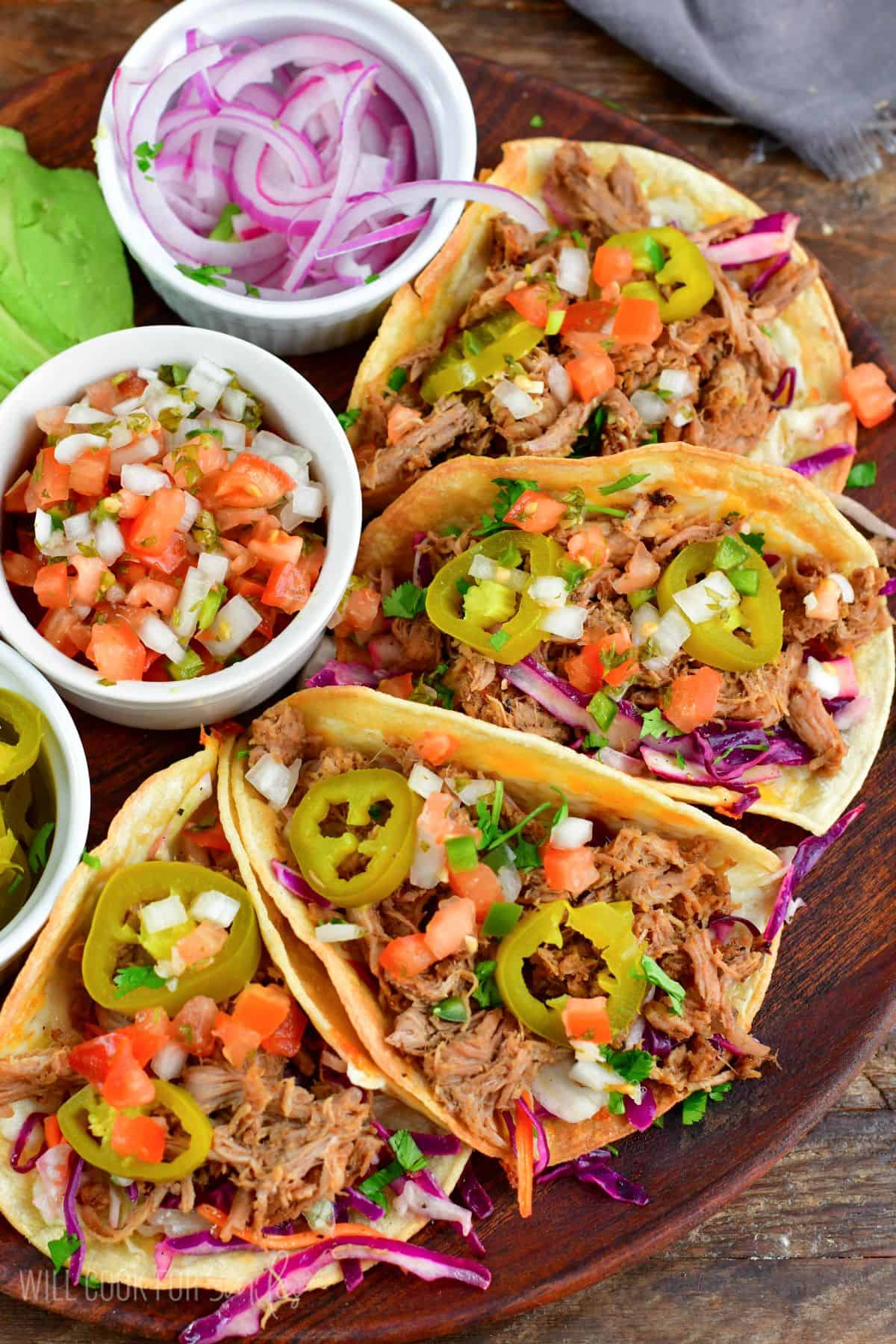 Four pulled pork tacos on a wooden plate with red onion, Pico de Gallo and pickled jalapenos in small bowls.
