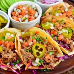 four pulled pork tacos on a wood plate with Tomato. Pico de Gallo and pickled jalapenos and small bowls with toppings.