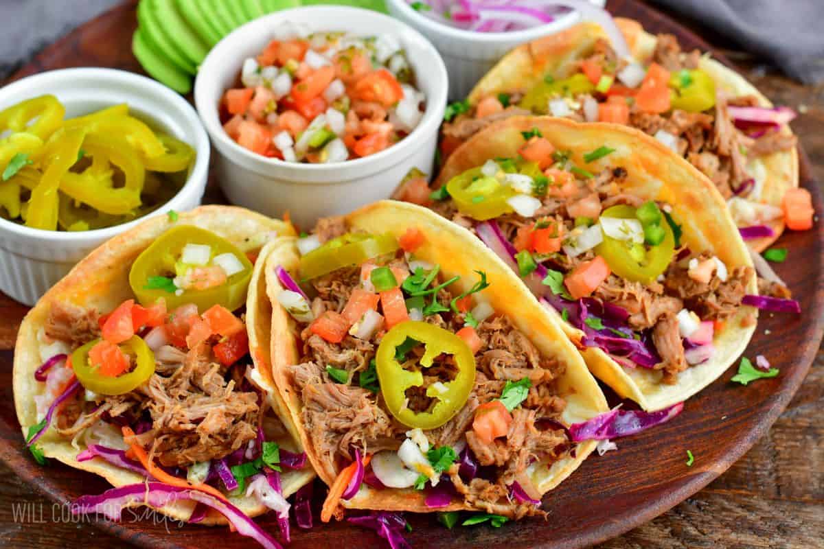four pulled pork tacos on a wood plate with Tomato. Pico de Gallo and pickled jalapenos and small bowls with toppings.