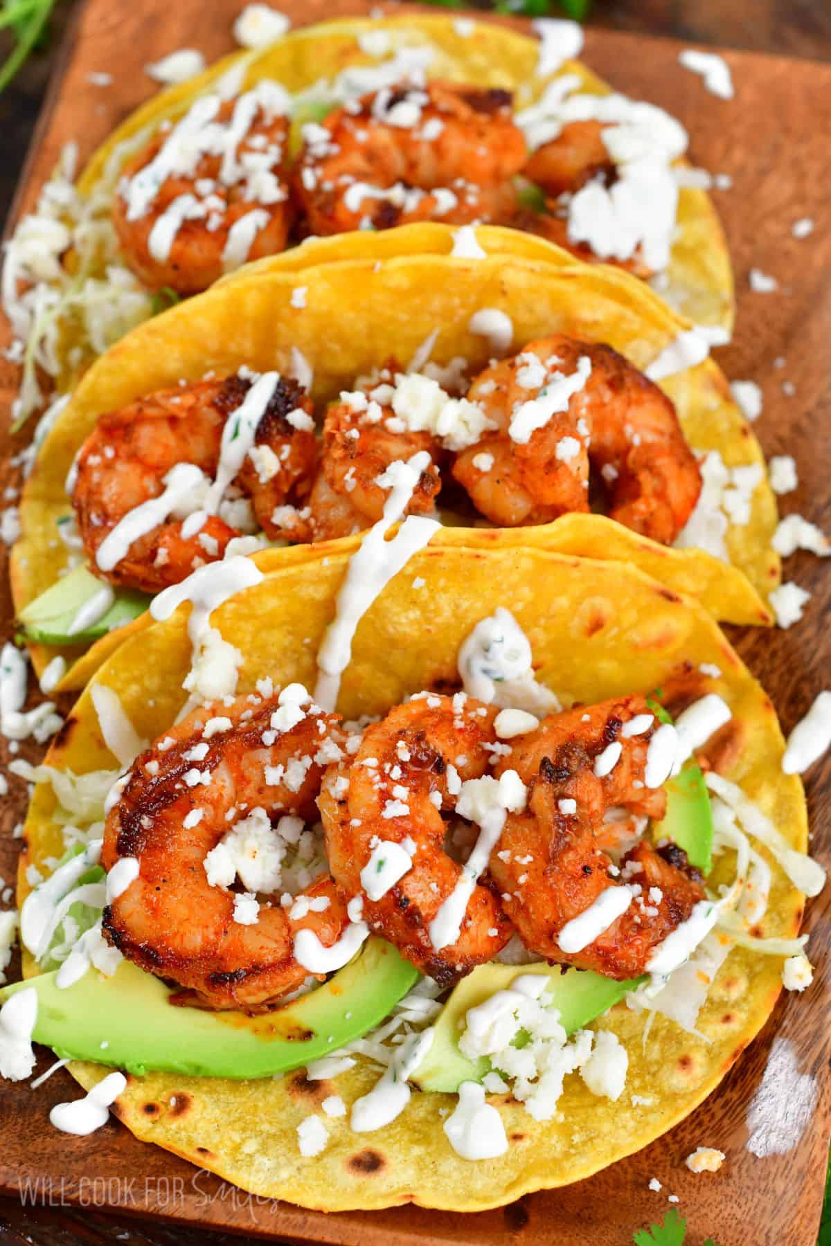 Three shrimp tacos on a wood plate with creme, avocado, an dcheese.