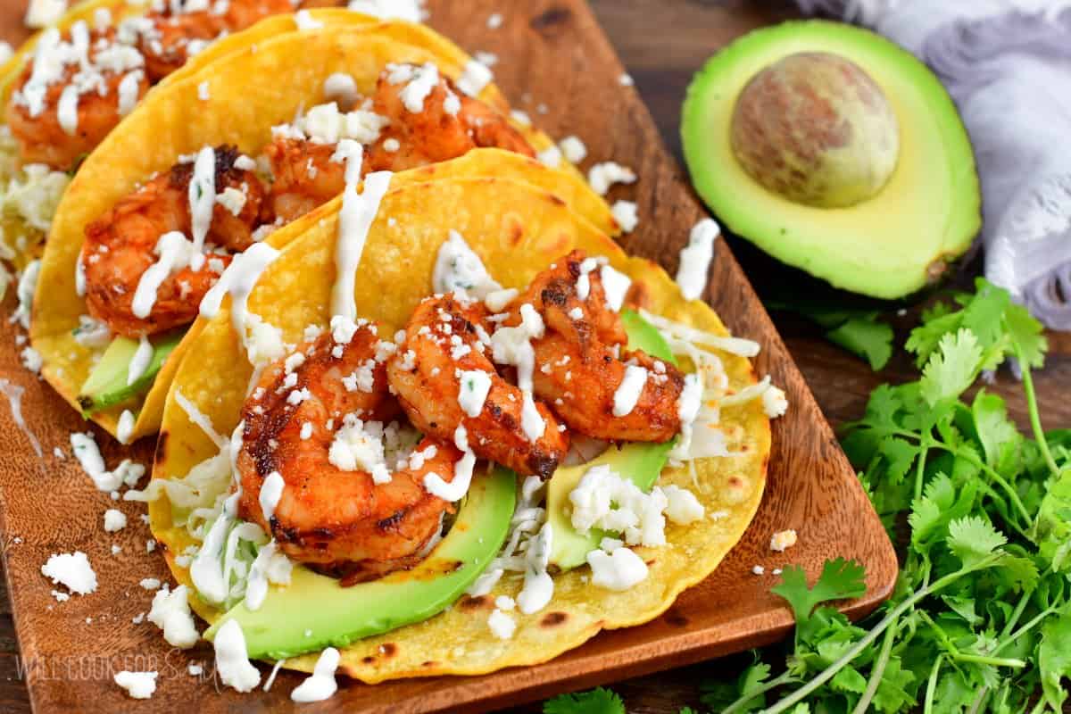 Shrimp tacos on a plate with cilantro and half an avocado on a wood surface.