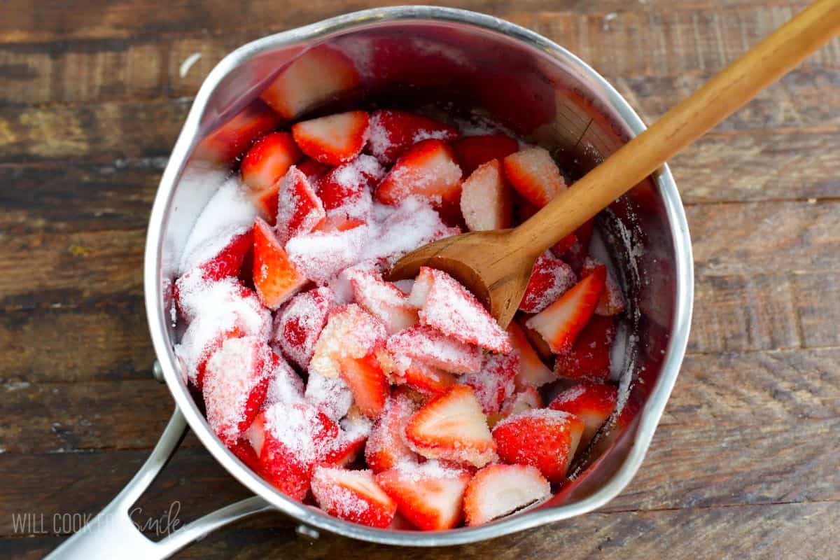 Strawberries in a pan with sugar over them and a wooden spoon.
