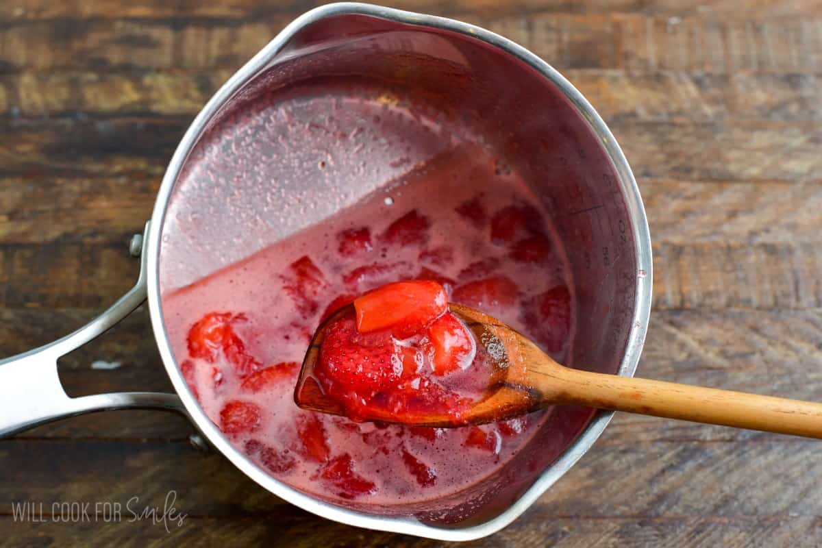 Cooked strawberries in a pan with some being scooped out on a wooden spoon.