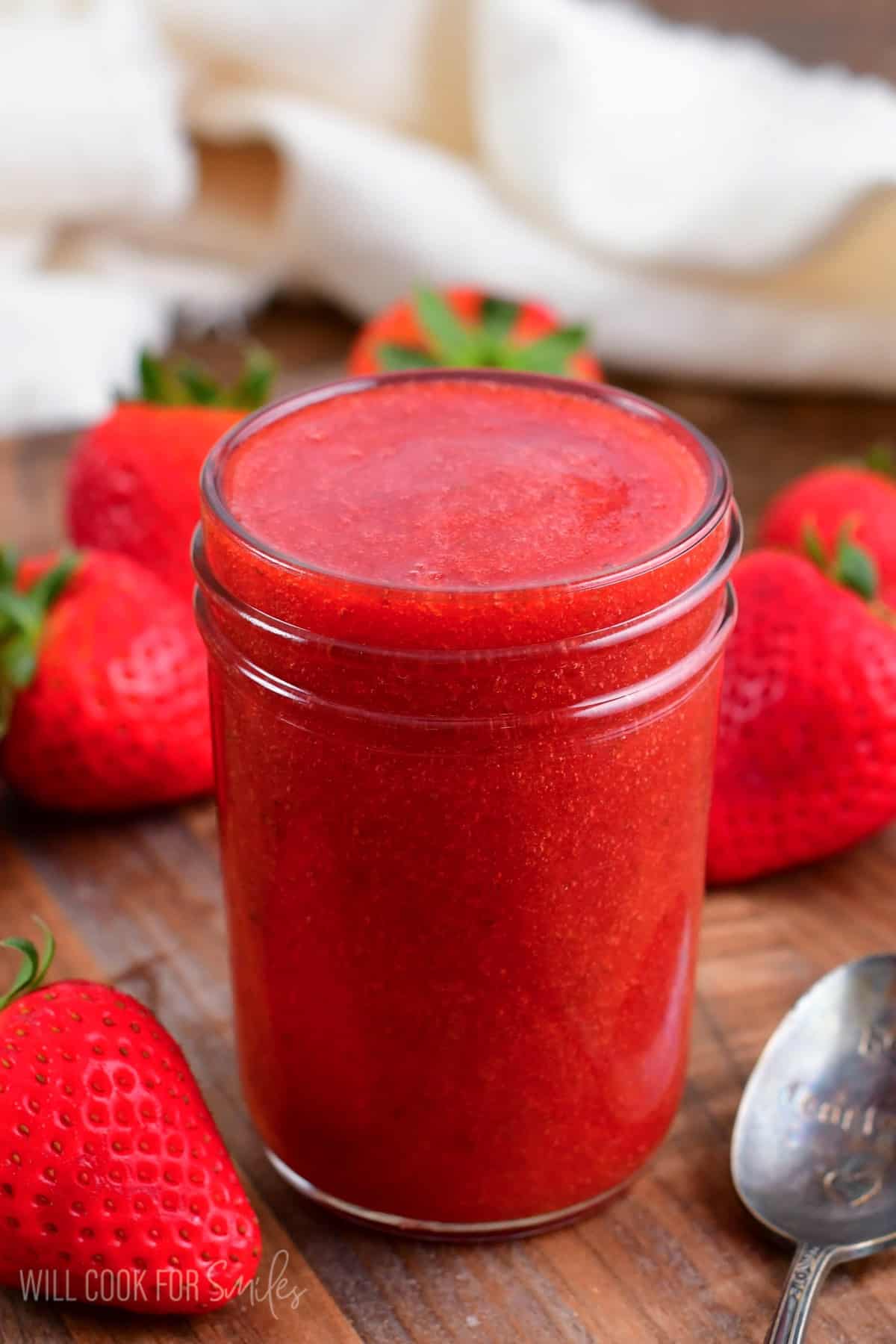 strawberry sauce in a glass jar on a wood surface with strawberries around it.