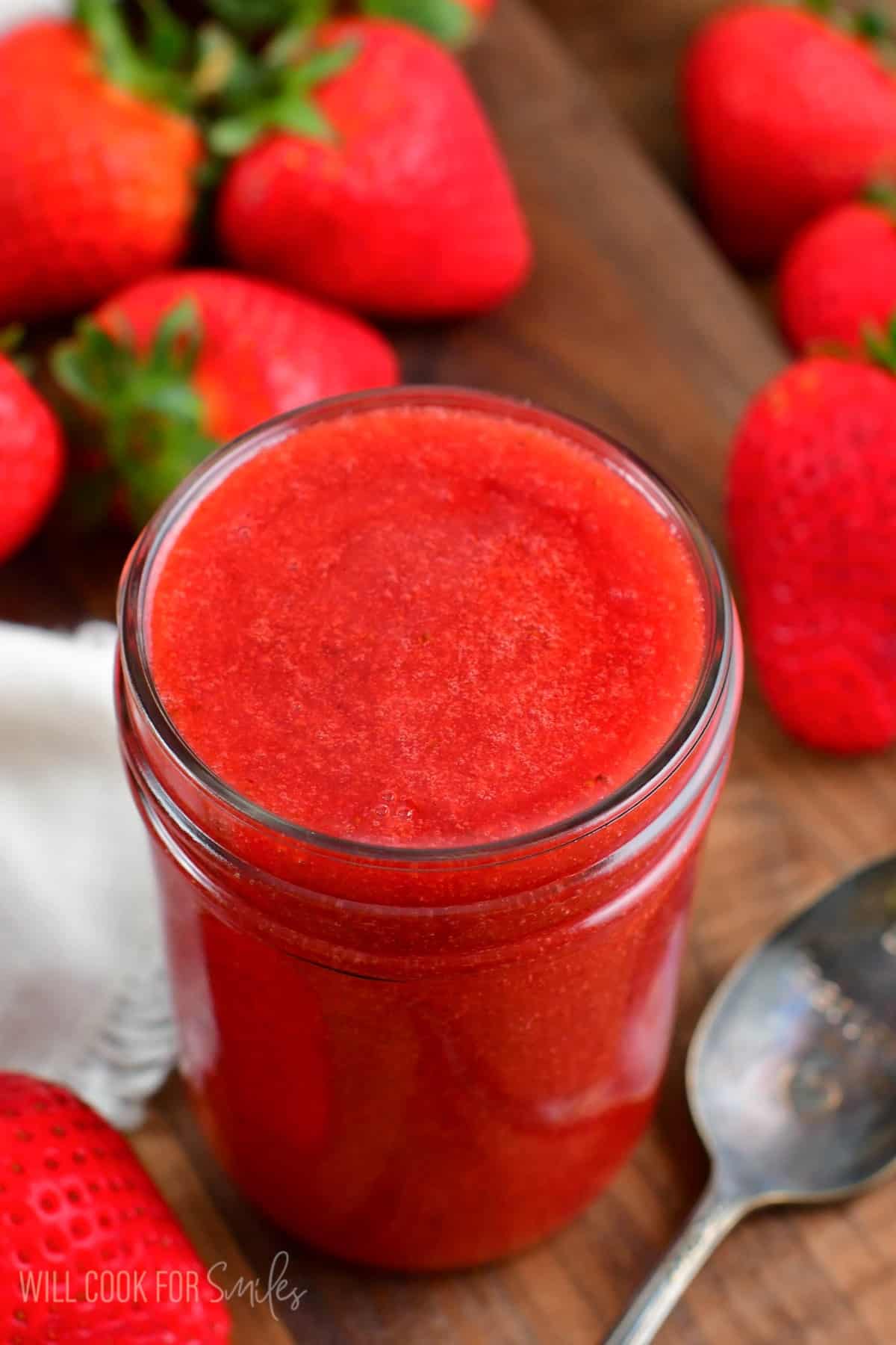Strawberry sauce in a mason jar with a spoon to the right and strawberries all around it.