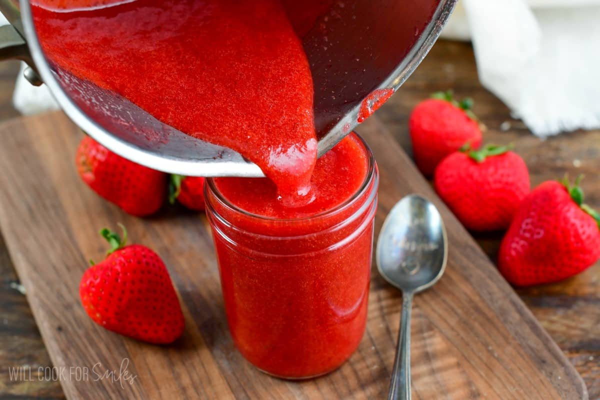 pouring strawberry sauce from the pot into a mason jar that is sitting on a wood cutting board.