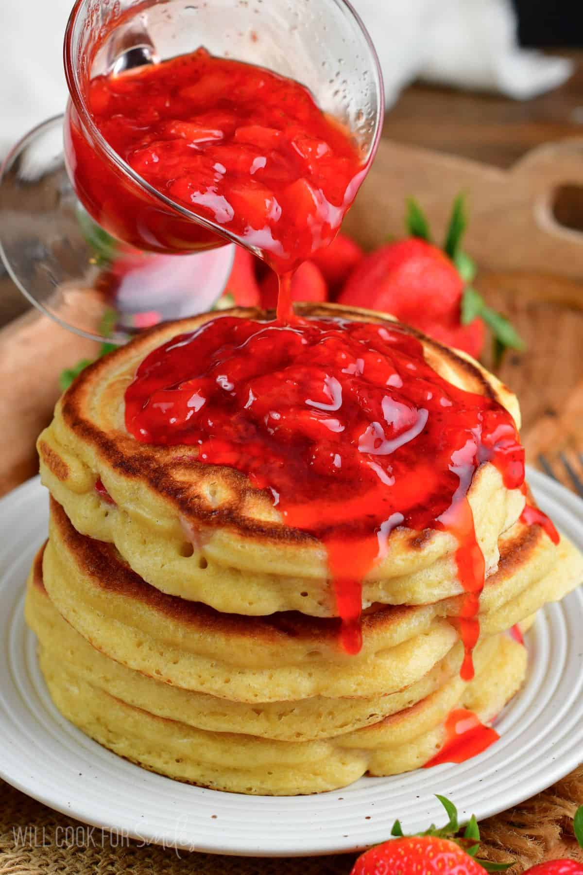 pouring strawberry topping over a stack of pancakes that are on a plate.