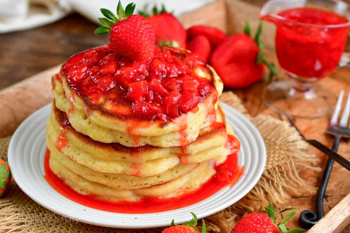 a stack of strawberry pancakes on a plate with strawberry topping and a whole strawberry on top.