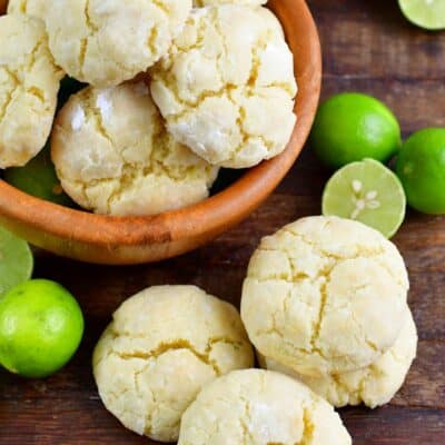 Cream cheese Cookies stacked in a wood bowl with a stack of cookies and limes beside the bowl on a wood surface.
