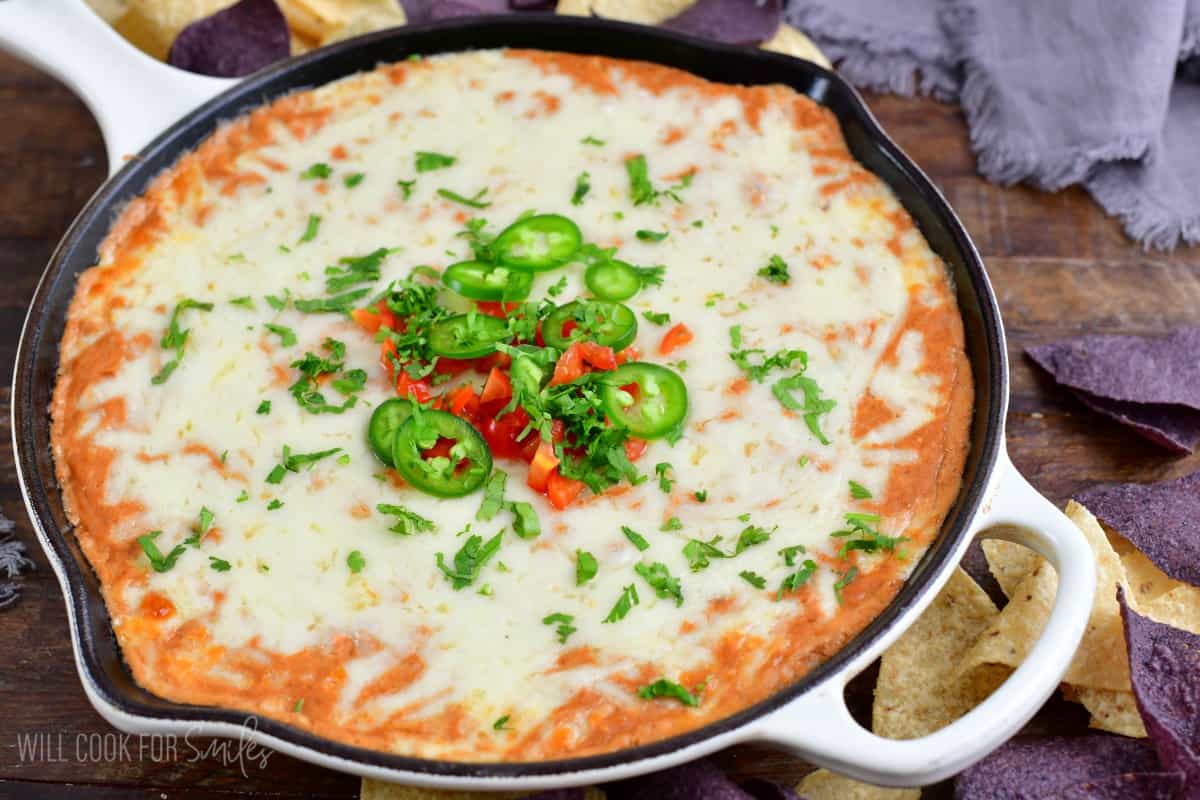 hot bean dip topped with melted cheese, tomatoes, jalapenos, and cilantro in a white cast iron skillet.