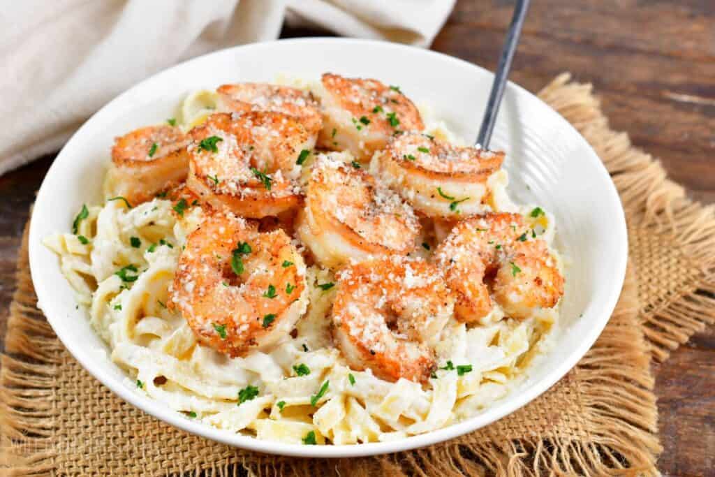A white bowl filled with creamy fettuccine pasta topped with grilled shrimp, sprinkled with grated cheese and chopped parsley. A fork rests on the side. The bowl is placed on a wooden table with a piece of burlap underneath.