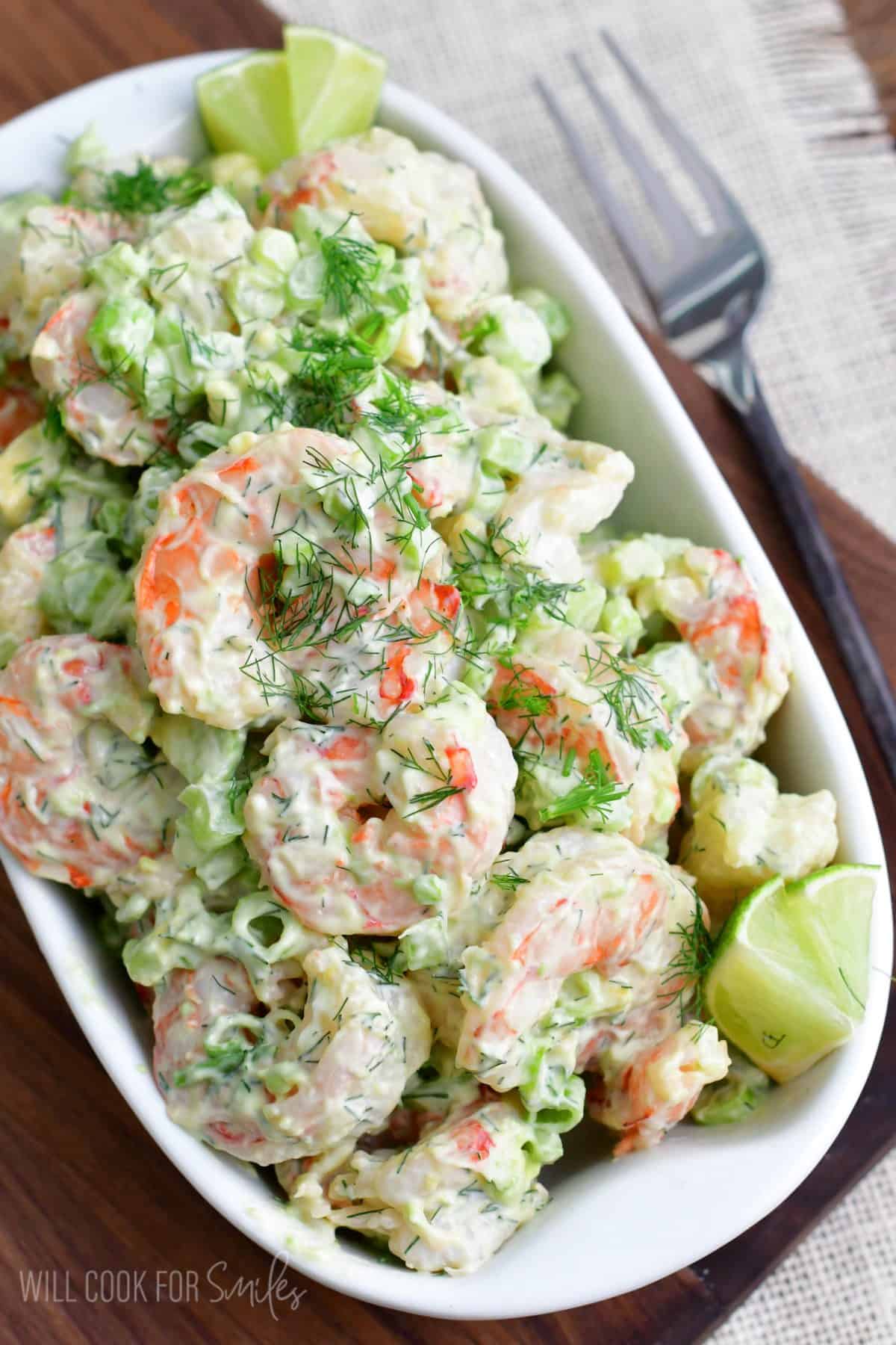 Shrimp salad in a dish topped with dill and garnished with lime wedges,.