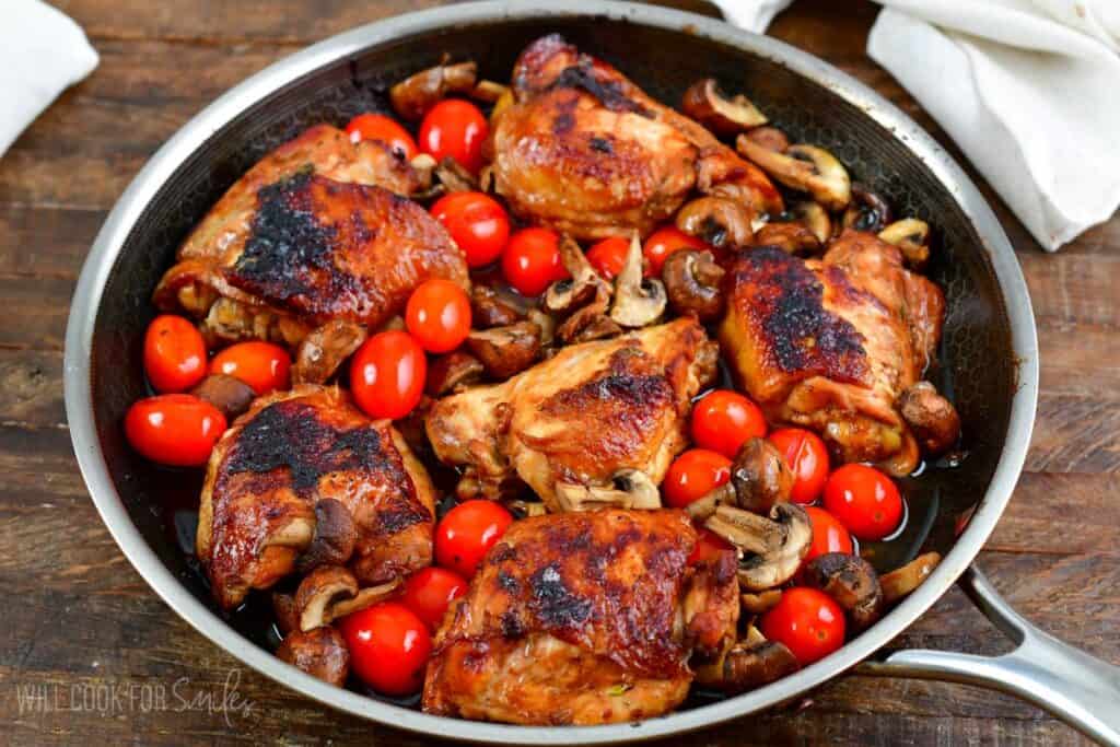 A skillet filled with six browned chicken thighs, surrounded by saut&eacute;ed mushrooms and cherry tomatoes. The caramelized chicken has a crispy exterior, and the vegetables add a burst of color with their red and brown hues. A white cloth is visible in the background.