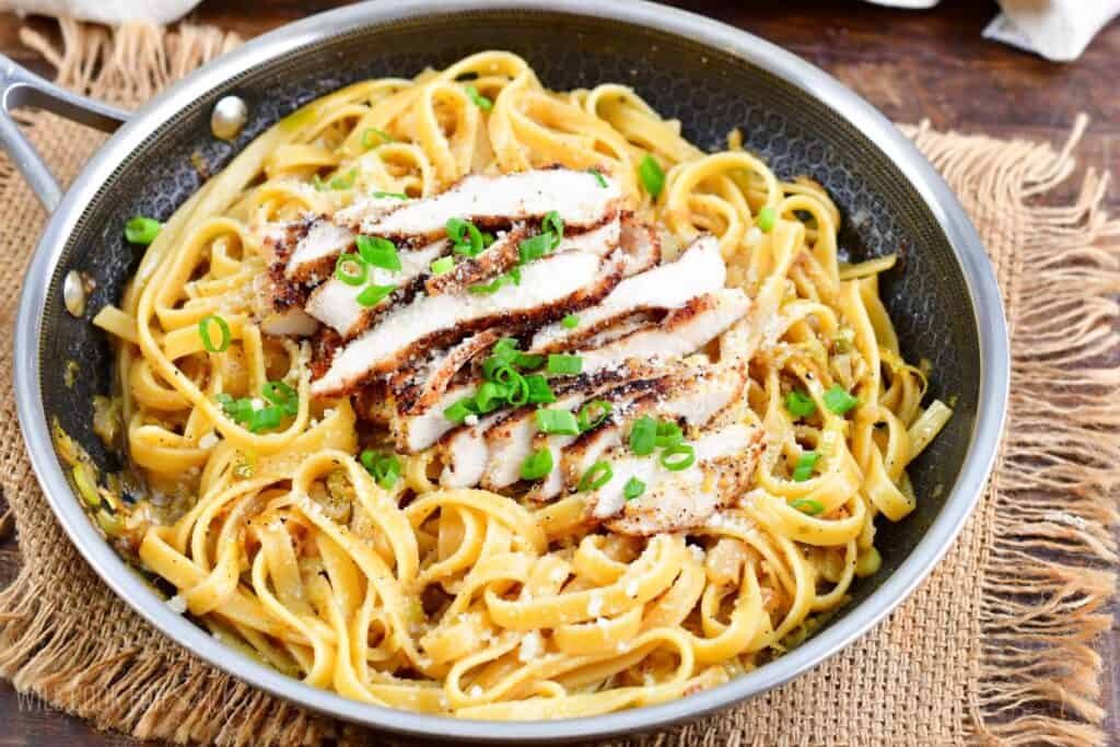 A skillet filled with creamy fettuccine pasta topped with slices of grilled chicken breast. The dish is garnished with chopped green onions and grated Parmesan cheese. The skillet rests on a burlap cloth on a wooden table.