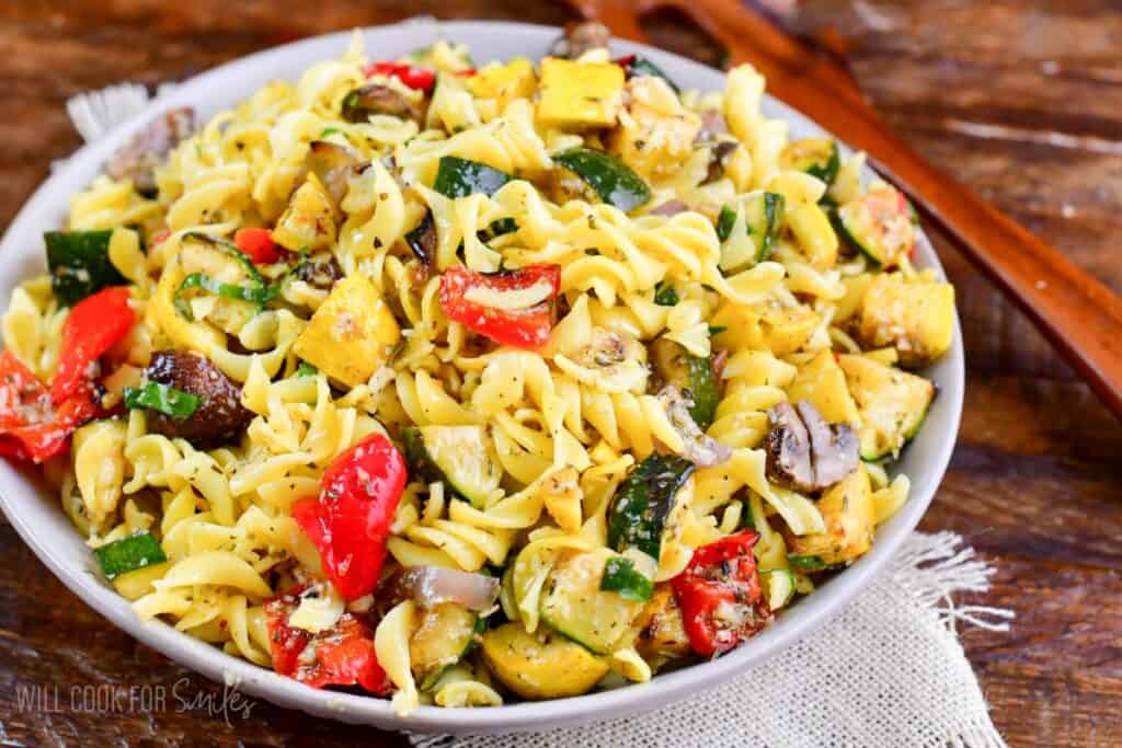 A white bowl filled with a colorful pasta salad featuring fusilli, grilled zucchini, red bell peppers, mushrooms, and sprinkled herbs. The bowl is placed on a wooden table with wooden serving utensils in the background.