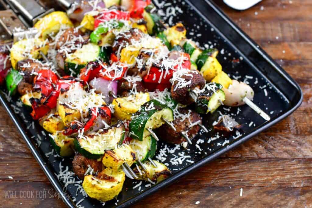 A tray of freshly grilled vegetable skewers featuring red bell peppers, zucchini, yellow squash, mushrooms, and onions, all sprinkled with grated Parmesan cheese. The vegetables are vibrant and charred, ready to be served.