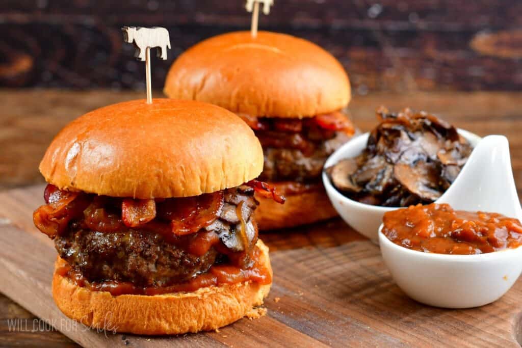 Two juicy burgers with melted cheese, crispy bacon, and saut&eacute;ed mushrooms are served on toasted buns. Each burger is held together with a skewer topped with a cow-shaped decoration. There are small dishes of saut&eacute;ed mushrooms and smoky barbecue sauce on the side.
