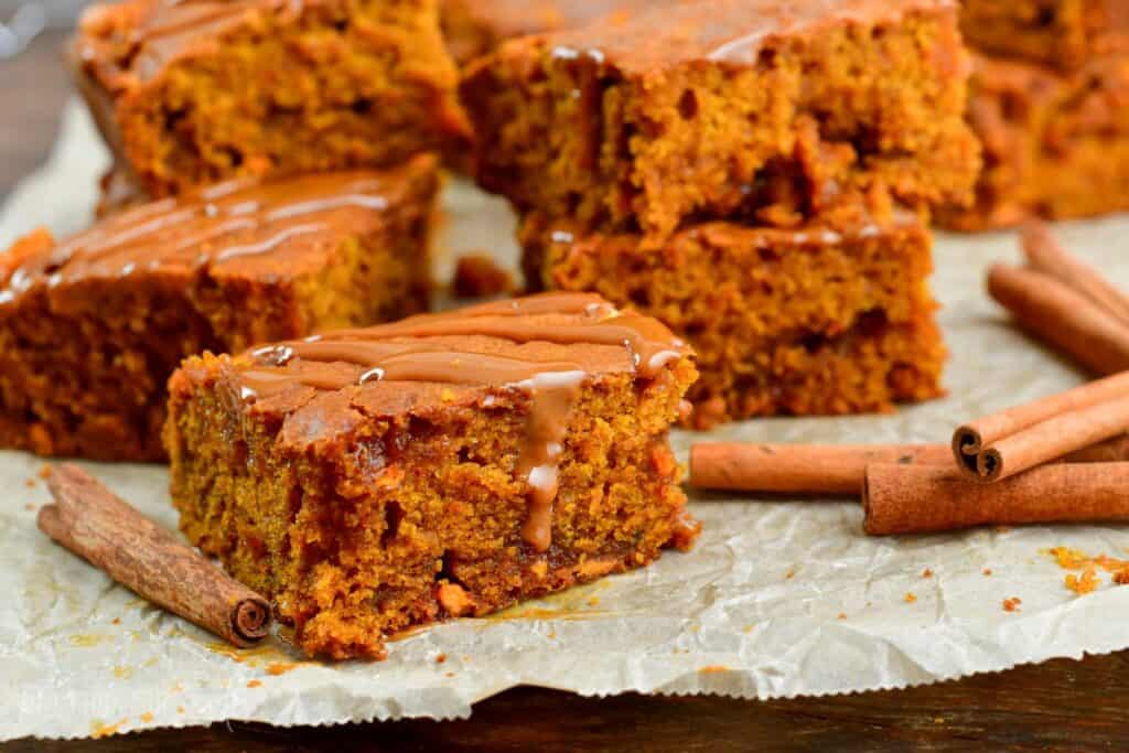 A close-up of pumpkin bars arranged on a parchment paper surface. The bars are moist and spiced, with a light drizzle of icing on top. Cinnamon sticks are scattered around, enhancing the autumnal feel of the scene. The bars have a rich, golden-brown color.