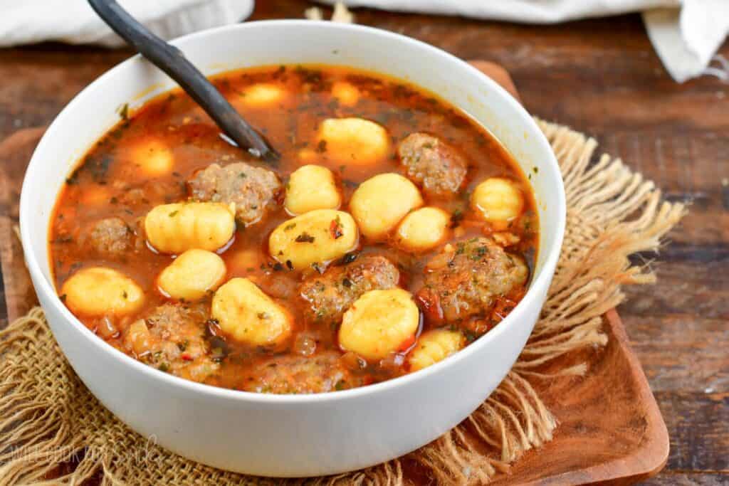 A white bowl filled with a hearty soup containing meatballs and gnocchi, garnished with herbs. A spoon rests inside the bowl, which is placed on a rustic wooden surface with a piece of burlap fabric underneath.