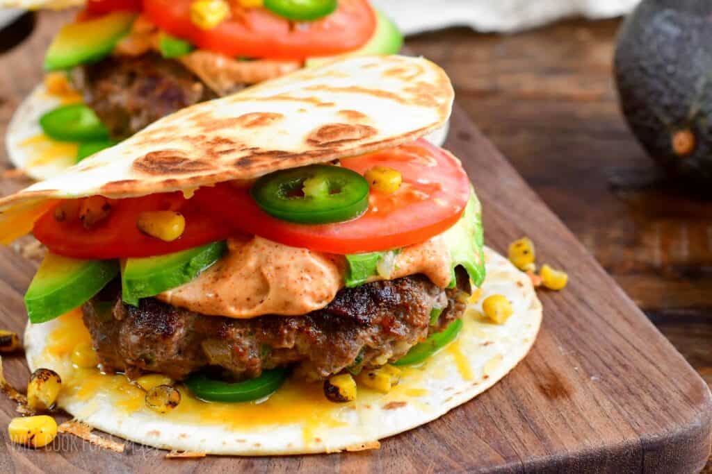 A vibrant taco is filled with a juicy grilled burger patty, sliced avocado, tomato, jalape&ntilde;o, corn, and a dollop of spicy sauce. The taco is lightly toasted and served on a wooden board with background details of an avocado and rustic surface.