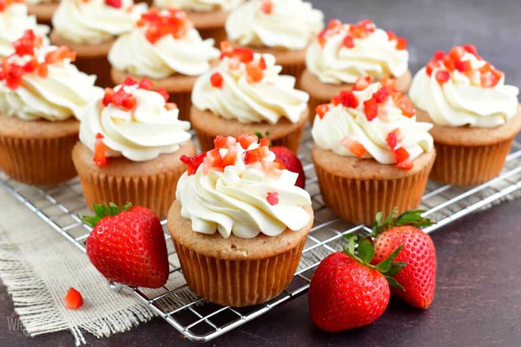 Close-up of strawberry cupcakes on a cooling rack. The cupcakes are topped with generous swirls of white frosting and sprinkled with small pieces of fresh strawberries. In the foreground, whole strawberries are visible, adding to the fresh, summery feel.