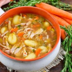 a bowl filled with clear chicken gnocchi soup with veggies and rosemary around.