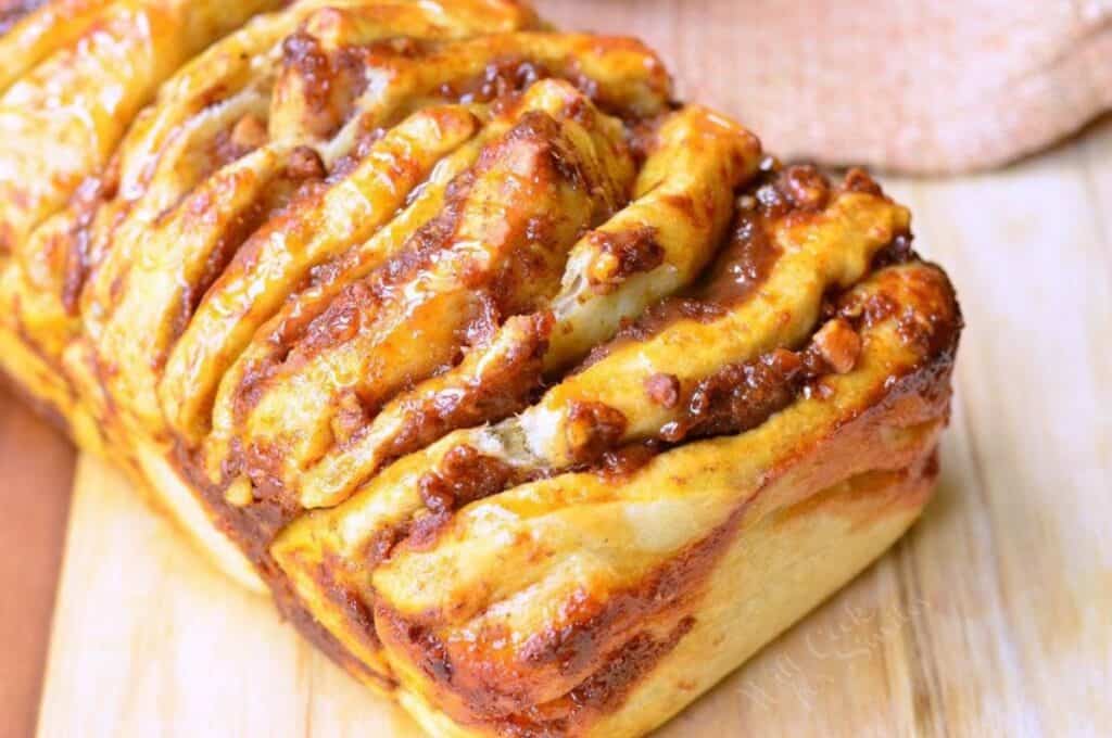 A close-up of a freshly baked loaf of cinnamon swirl bread on a wooden cutting board. The bread has a golden-brown crust with visible swirls of cinnamon and sugar, creating a glossy, caramelized texture on the surface.