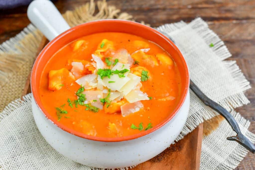 A white bowl filled with creamy tomato tortellini soup, topped with shaved parmesan and chopped parsley, placed on a rustic wooden surface with a burlap cloth underneath.