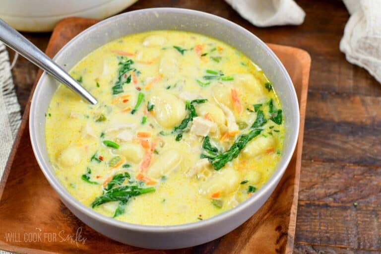 a bowl full of creamy chicken gnocchi soup with a spoon and sitting on a wooden plate.