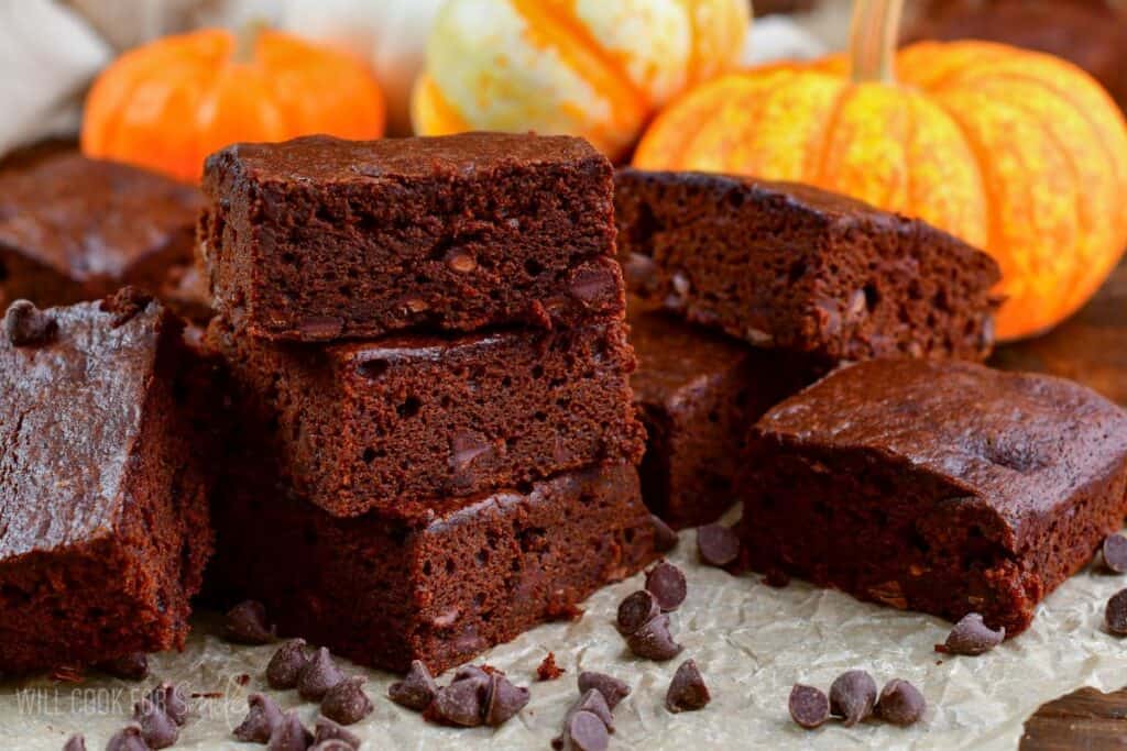 A stack of chocolate brownies surrounded by scattered chocolate chips on parchment paper. In the background, small pumpkins and gourds add a festive autumn touch.