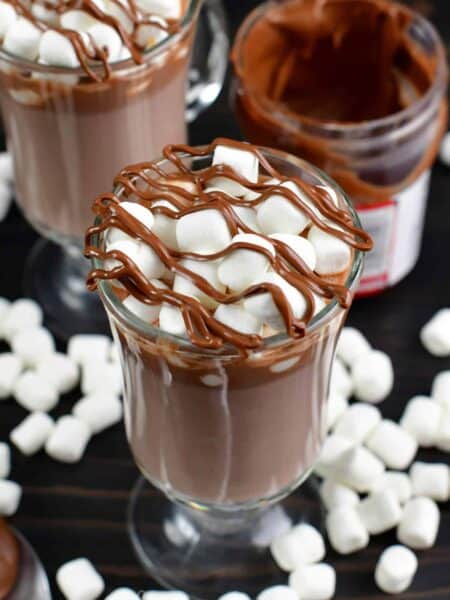 Nutella hot chocolate in a glass with marshmallows on a wood surface with marshmallows around the bottom.