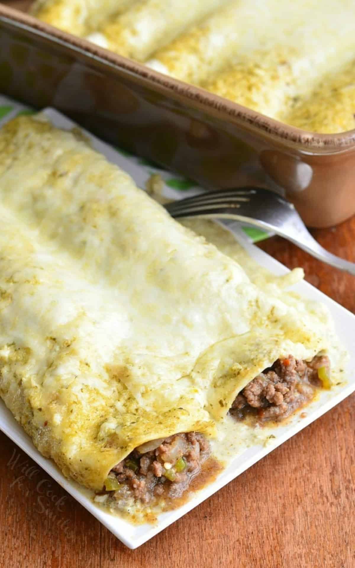 two beef enchiladas on the plate next to the baking dish with more enchiladas.