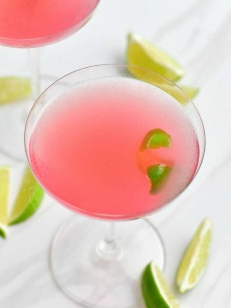 light pink cosmopolitan cocktail in a martini glass with lime wedges around.