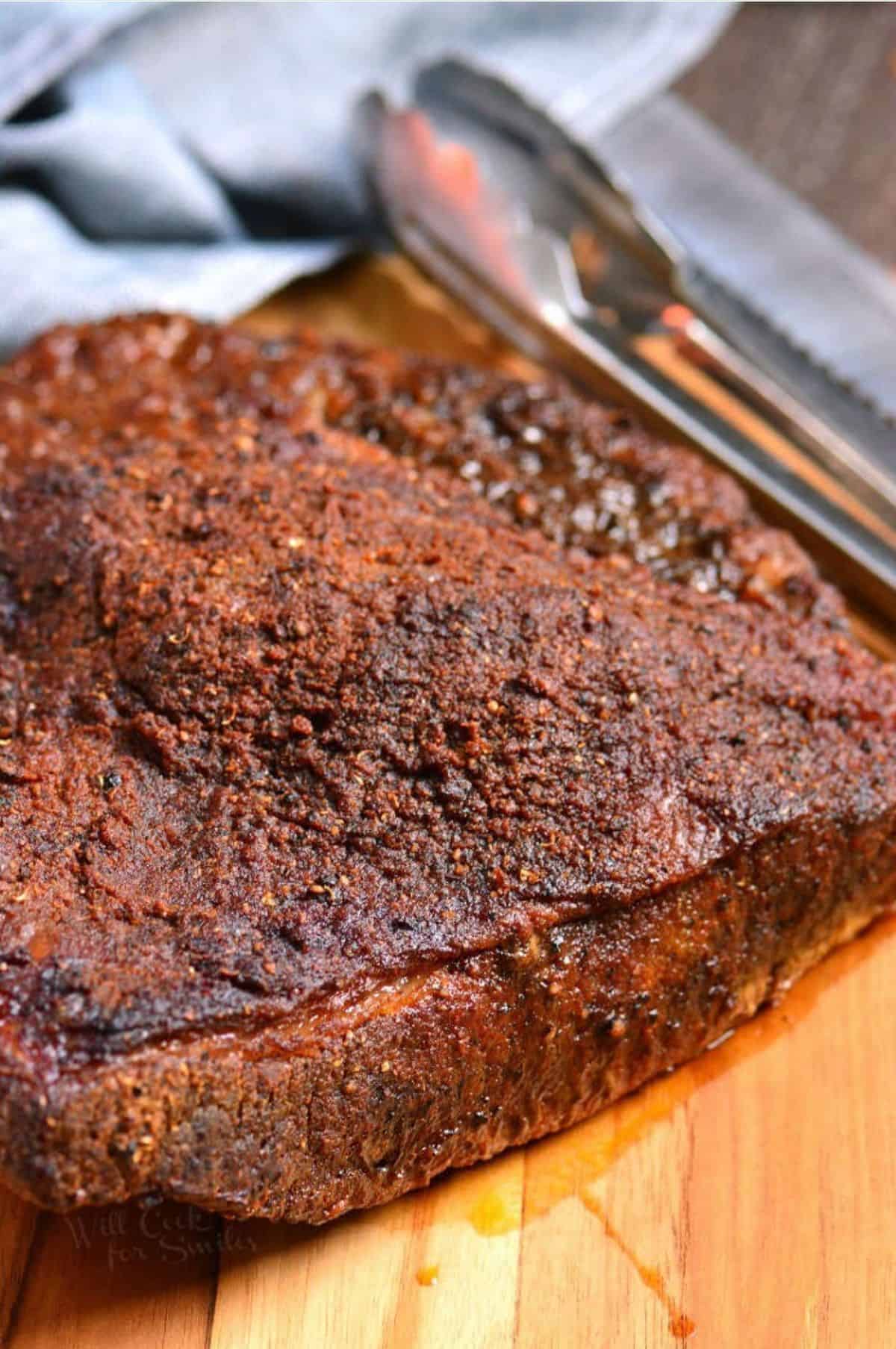 Cooked beef brisket resting on the cutting board with tongs and a knife next to it.
