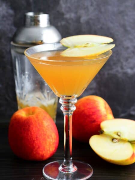 apple martini in a martini glass with a slice of apple on top as garnish with 3 apples one sliced on table.