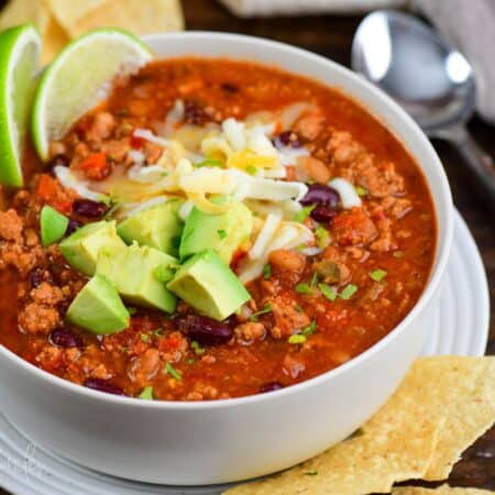 turkey chili in a bowl topped with cheese, avocado, lime wedges, and cilantro.