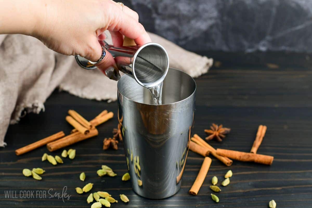 adding vodka to cocktail shaker with cinnamon sticks and cardamom seeds around the bottom.
