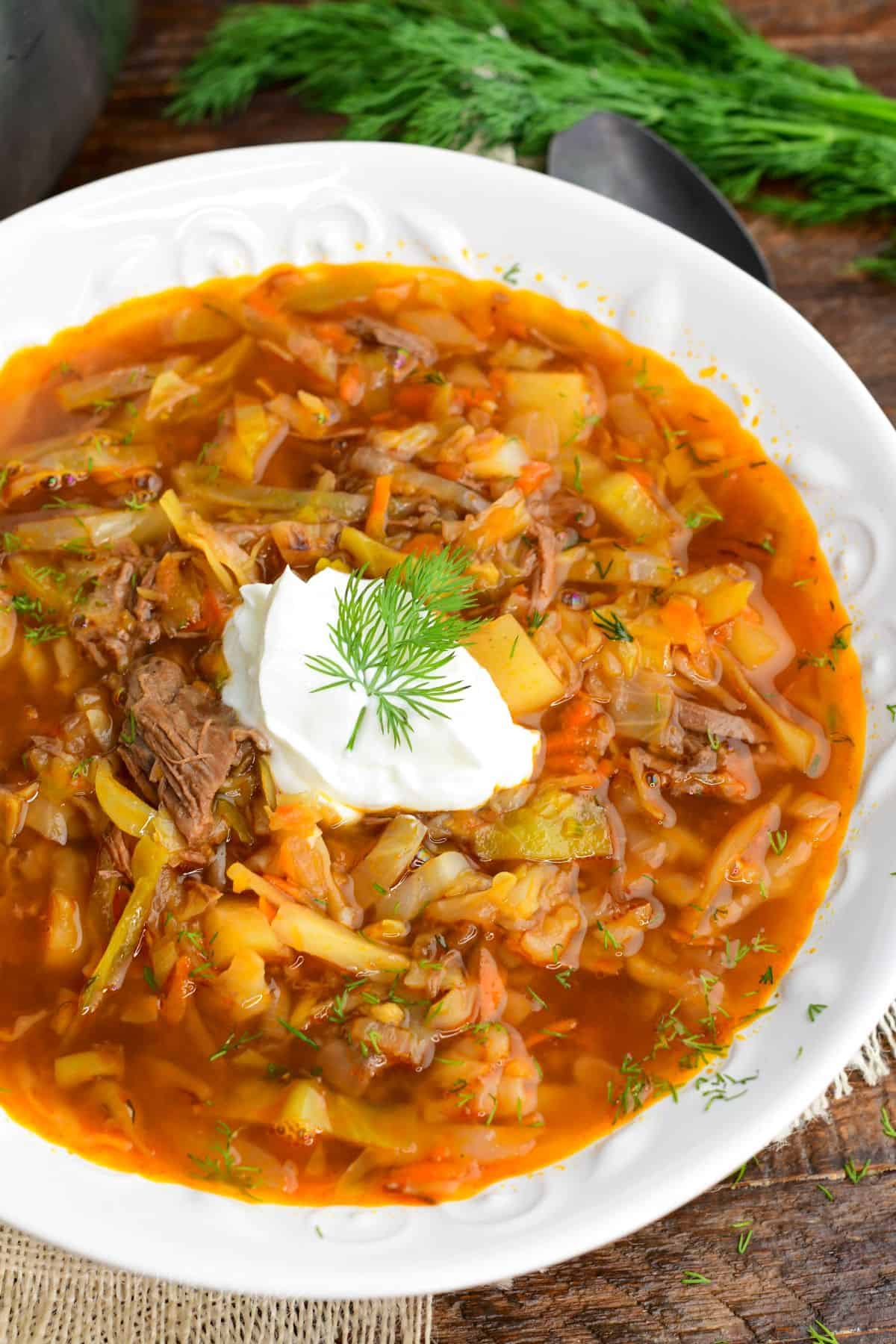 Russian beef and cabbage soup (shchi) in a white bowl with sour cream and dill and a spoon next to it.
