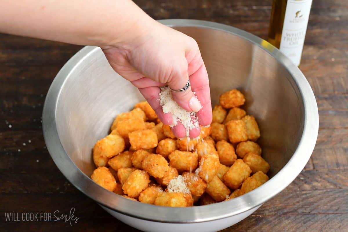 Adding parmesan cheese to a metal bowl of cooked tater tots.