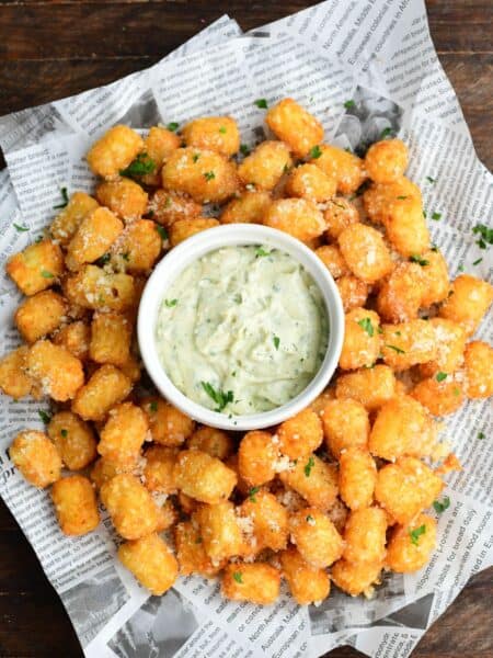Truffle tater tots on a lined tray with a garlic herb aioli in a bowl in the middle on a wood surface.