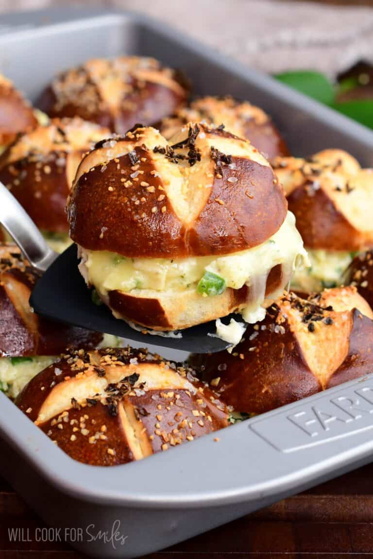 A baked jalapeño ranch chicken slider on a pretzel bun being lifted from the pan, showing melted cheese and creamy chicken filling.