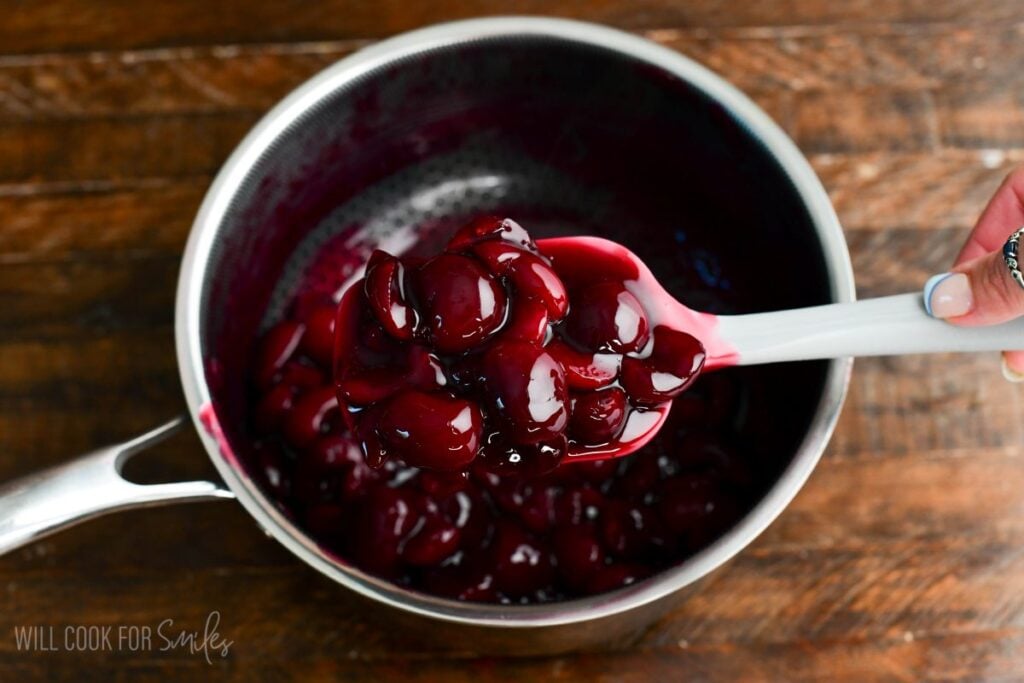 Cooked cherry topping in a saucepan being stirred with a spatula for no-bake cherry cheesecake parfaits.