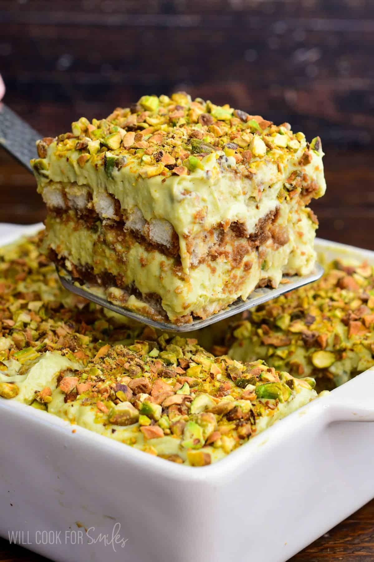 Close-up of a thick slice of pistachio tiramisu being lifted from a white baking dish, showing creamy pistachio layers, soaked ladyfingers, and a crunchy chopped pistachio topping.