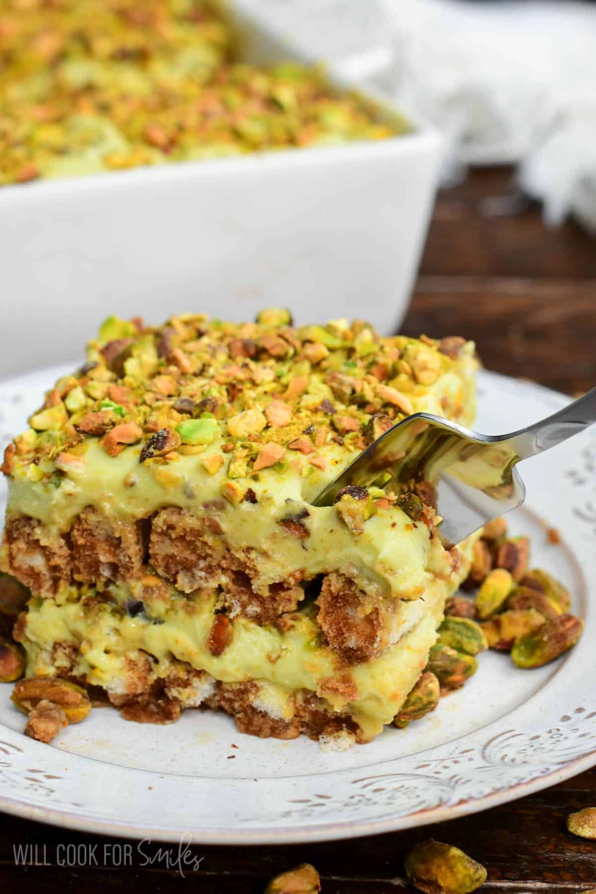 Fork cutting into a creamy slice of pistachio tiramisu topped with chopped pistachios, showing layers of pistachio mascarpone filling and coffee-soaked ladyfingers on a plate.