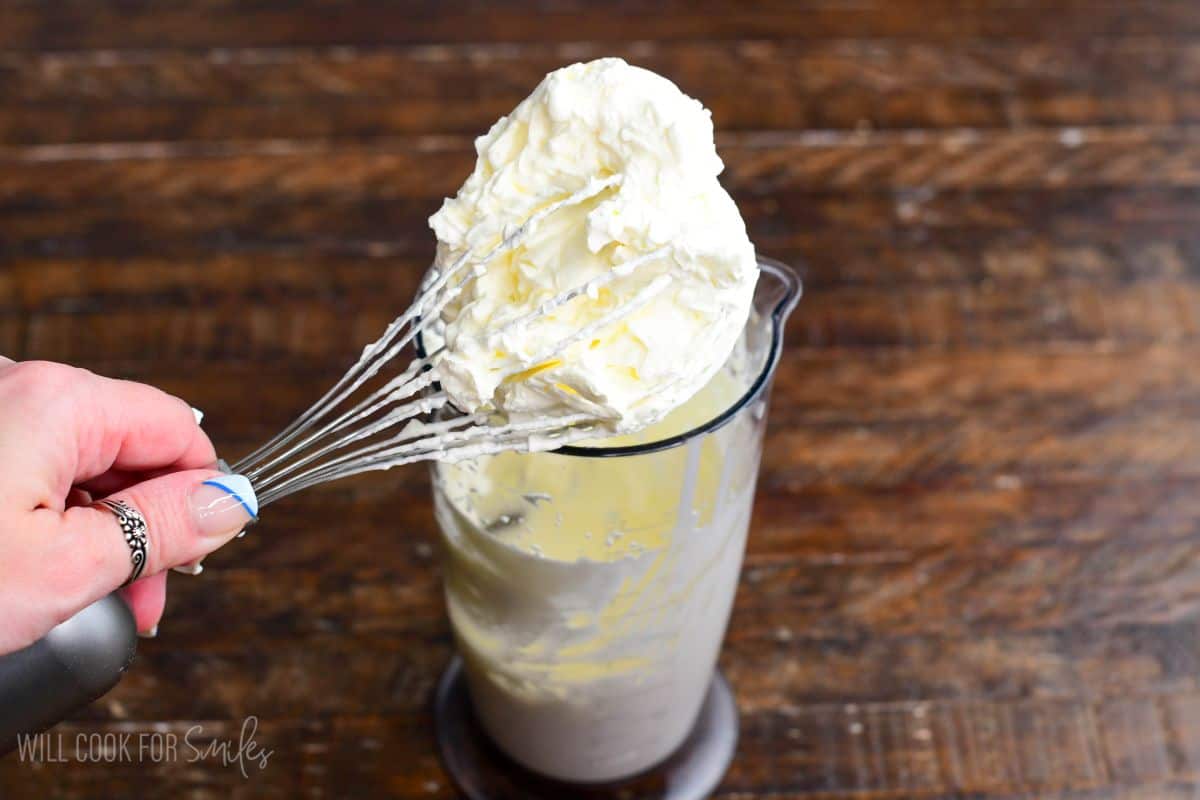 Fresh whipped cream being lifted from a mixing cup with a whisk, showing thick soft peaks.