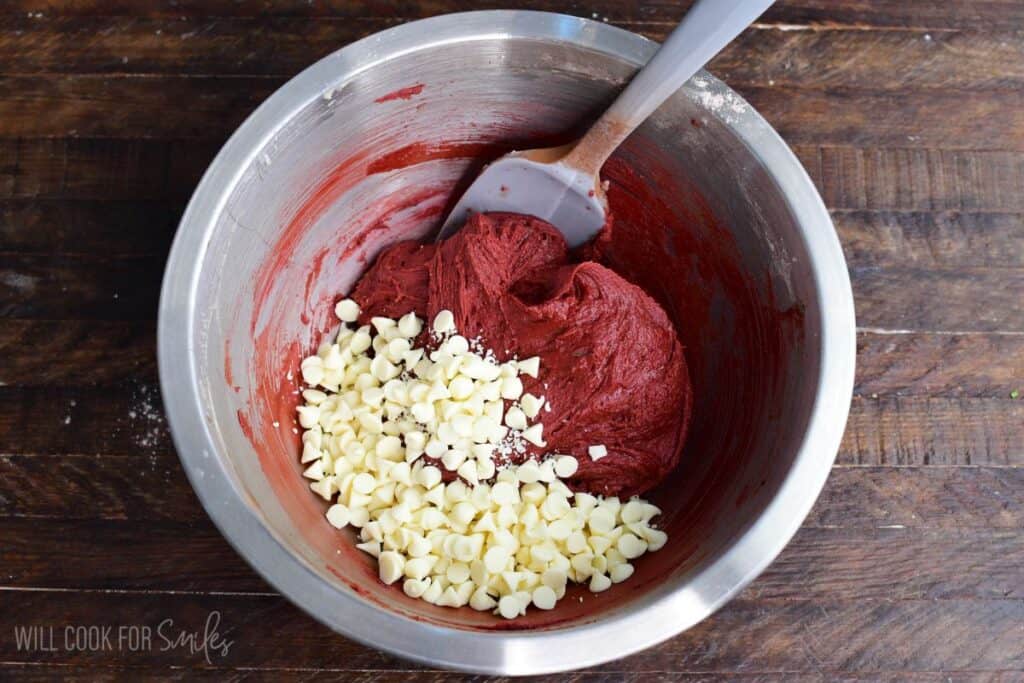White chocolate chips added to red velvet cookie dough in a mixing bowl for red velvet cream cheese cookies.