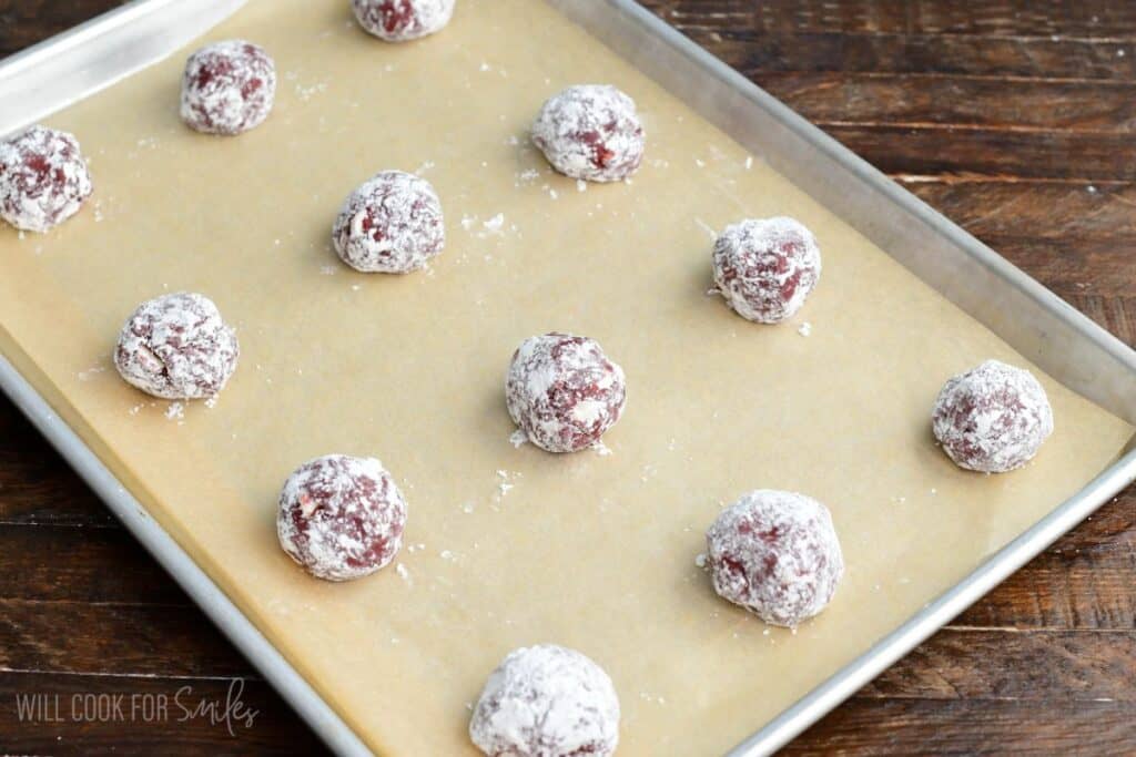 Red velvet cream cheese cookie dough balls coated in powdered sugar arranged on a parchment lined baking sheet before baking.