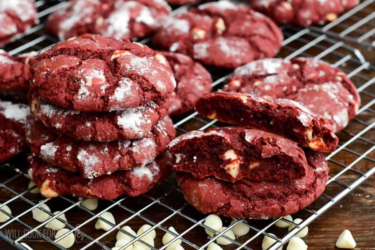 Red velvet cream cheese cookies stacked on a wire rack with one cookie broken open to show the soft center and white chocolate chips.